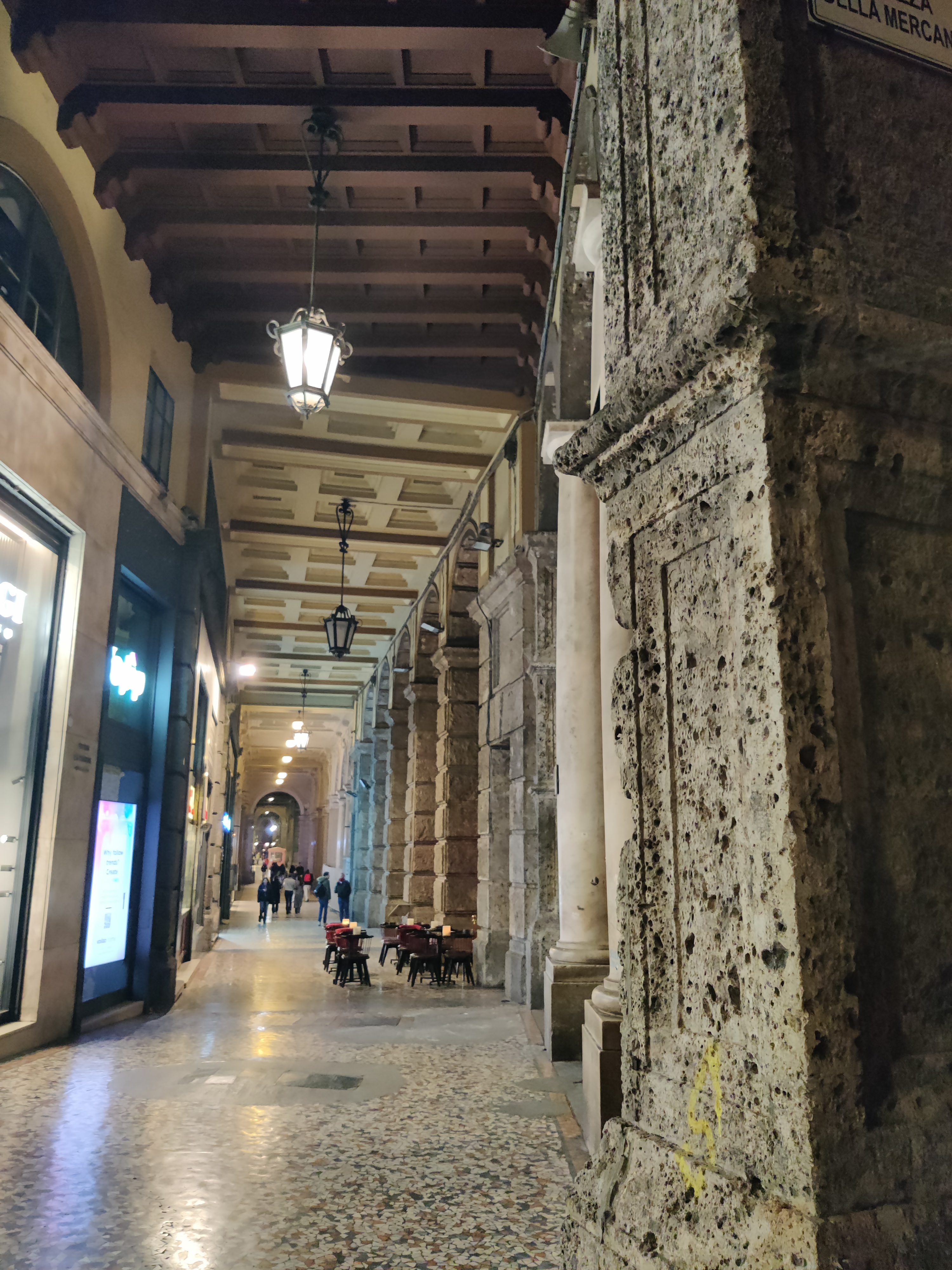 Night-time Bologna portico with worn stone columns, hanging lanterns and a street sign for Piazza della Mercanzia